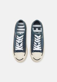 Matala siniset farkkikengät valkoisilla nauhoilla ja vaaleanbeigeillä kumikärjillä, nähtynä ylhäältä valkoisella taustalla.