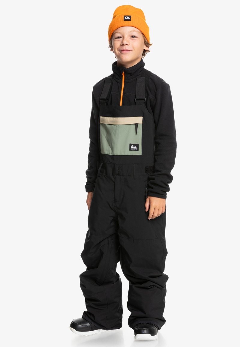 Svart snöoverall med justerbara ремmar, beige och grön bröstficka, tillsammans med en svart zip-up topp och en orange mössa. Svarta kängor.