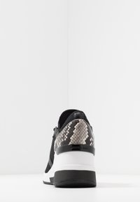 Zapatilla negra con talón de patrón de piel de serpiente, media suela blanca y parte superior texturizada. Presenta cierre de cordones y suela en contraste.