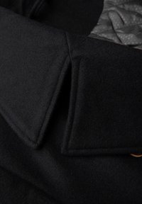 Col de manteau en laine noir avec des bords tranchants, présentant un intérieur matelassé en tissu gris. La texture est douce, avec des coutures visibles et des accents.