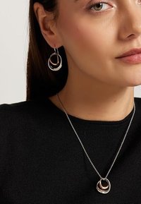 Collar de tono plateado con un colgante de doble lazo, acompañado de pendientes de aro a juego con acentos dorados. Usado sobre un suéter negro.