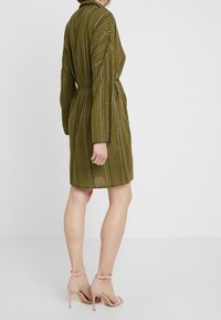 Robe verte olive, longueur genou, avec des rayures verticales et une taille cintrée. Manches longues et texture douce. Assortie à des escarpins rose clair.