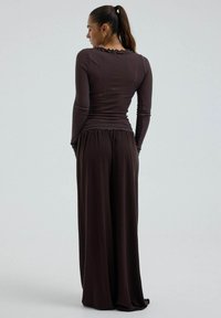 Femme avec une queue de cheval portant un haut ajusté à manches longues et un pantalon à jambes larges, debout de dos face au spectateur sur un fond uni.