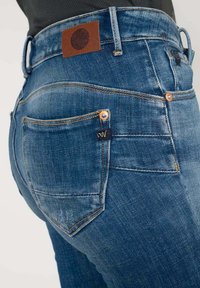 Jean en denim bleu au design ajusté, avec des coutures orange, une étiquette en cuir marron sur la ceinture et des poches arrière avec une marque.