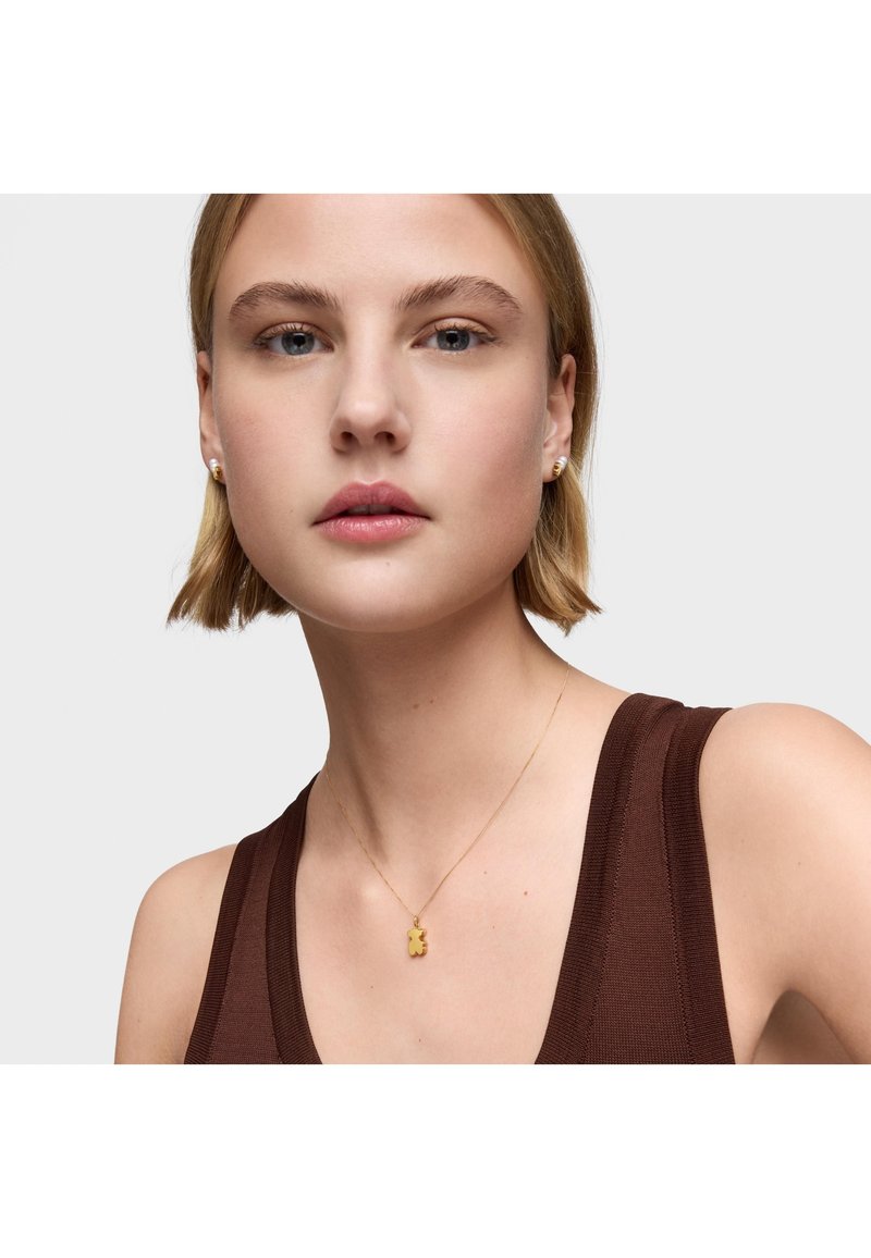 Collana con pendente a forma di orso in oro su una catena sottile, indossata sopra una maglietta senza maniche marrone, con una superficie metallica liscia e un design minimalista.