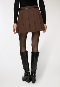 Gonna mini plissettata marrone con cintura nera, abbinata a stivali neri con tacco alti fino al ginocchio. La gonna presenta un tessuto testurizzato e una forma strutturata.