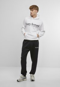 White hoodie with front pocket and black logo text paired with black sweatpants. Both garments feature a relaxed fit and elastic waistband.