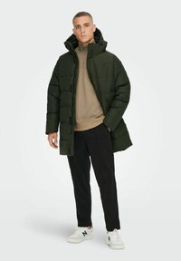 Doudoune verte avec capuche, présentant un design matelassé, un système de fermeture à boutons et des poches latérales, portée sur un haut beige à col zippé et un pantalon sombre.
