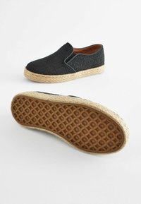 Ein Paar schwarze Slipper mit geflochtenen Textur-Oberteilen und geflochtenen Jutesohlen, wobei eine Sohle mit strukturiertem Gummi gezeigt wird.