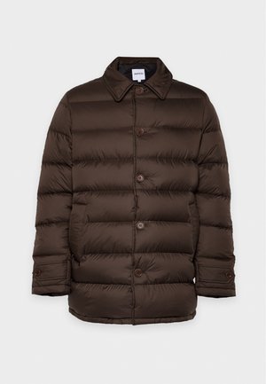 Un homme porte un manteau matelassé boutonné marron foncé, un pantalon noir et des bottes en cuir, sur un fond clair et uni.