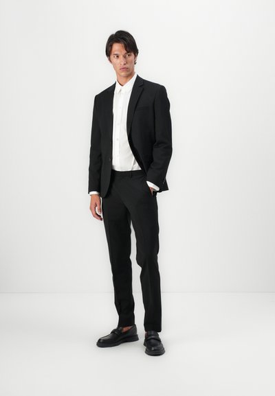 Costume noir ajusté avec une veste cintrée et un pantalon slim. Chemise blanche en dessous, assortie à des mocassins en cuir noir. Texture de tissu lisse.