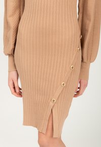 Strickkleid in Beige mit vertikalem Rippenmuster, seitlichem Schlitz und goldfarbenen Knopfakzenten entlang der linken Seite. Weiche Textur und figurschmeichelndes Design.