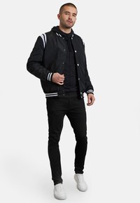Schwarze Bomberjacke mit weißen Streifen an den Ärmeln, Druckknöpfen, über einem schwarzen Hoodie getragen. Kombiniert mit schwarzen Hosen und weißen Sneakers.