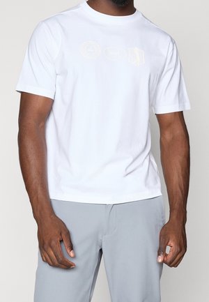Uomo che indossa una t-shirt bianca a maniche corte con loghi sfumati sul petto e pantaloni grigio chiaro, in piedi davanti a uno sfondo semplice.