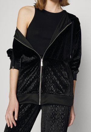 Femme portant un haut noir sans manches sous un sweat à capuche en velours noir partiellement dézippé et un pantalon assorti avec des lettres à motif subtil.