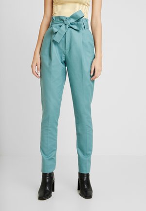 Mujer con pantalones paperbag de cintura alta color teal con cinturón anudado, blusa sin mangas amarilla con rayas, y botines negros con tacón, de pie.