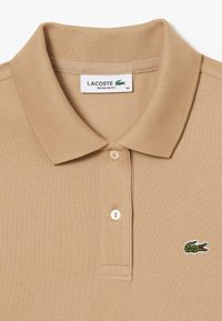 Beige polo paita, jossa on teksturoitu kangas, kolme nappia ja pieni kirjailtu vihreä krokotiililogo vasemmassa rinnassa. Normaalimitoitus.