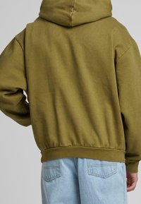 Olivově zelená mikina s klokaní kapsou, žebrovanými manžetami a spodním lemem. Měkká látka s uvolněným střihem, zobrazená zezadu. Viditelné světle modré džíny.
