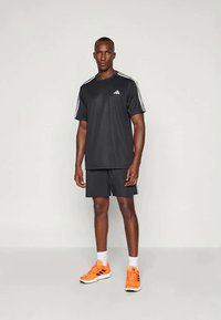 Svart sportsklærsett med kortermet T-skjorte og shorts med hvite striper. Modellen har på seg klare oransje joggesko.