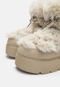 Beige platform csizmák műszőrme díszítéssel, lekerekített orral, vastag talppal és díszkötésekkel. Texturált felsőrész varrott részletekkel.