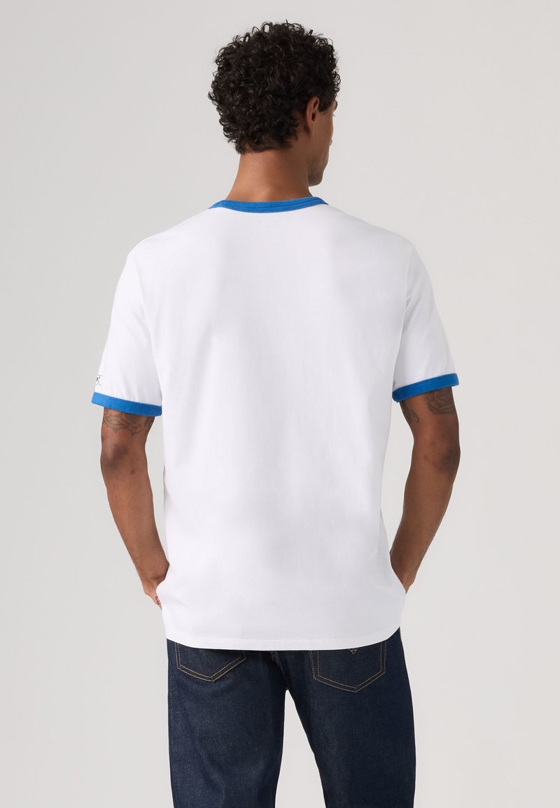 Homme aux cheveux bouclés, debout de dos, portant un t-shirt blanc avec un liseré bleu au col et aux manches et un jean bleu foncé.