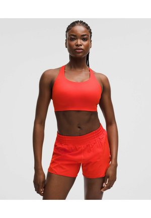 Athletische Frau in rotem Sport-BH und passenden Shorts steht selbstbewusst vor einem schlichten hellen Hintergrund.