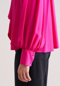 Blusa fucsia con maniche a palloncino, tessuto morbido e polsini decorati con bottoni, abbinata a pantaloni in denim neri.