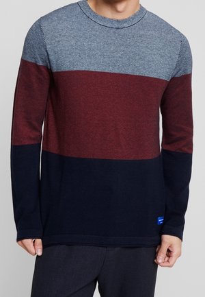 Homme portant un pull en maille à manches longues avec de larges rayures horizontales en gris clair, bordeaux et bleu marine.