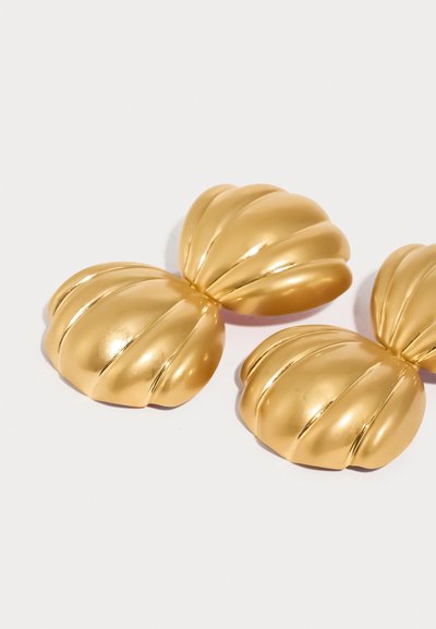 Διακοσμητικά κομμάτια σε χρυσό τόνο με σχεδιασμό που μοιάζει με ραβδώσεις και κοχύλι. Η λεία, αντανακλαστική επιφάνεια αναδεικνύει το τρισδιάστατο σχήμα τους.