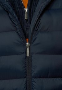 Marineblauwe pufferjack met doorstikte secties, ritssluiting en een oranje binnenlaag met een gestructureerd breisel ontwerp.