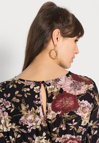Blusa floral preta com flores rosas e burgundy. Apresenta um detalhe em fenda nas costas e decote redondo. Brincos de argola dourados visíveis.
