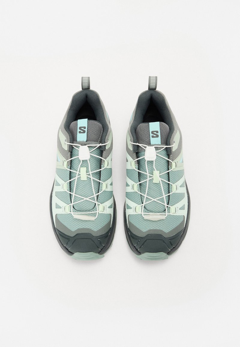 Des chaussures de trail à lacets avec un dessus en mesh vert clair et gris, une semelle en caoutchouc épaisse, des rainures bien marquées et des accents réfléchissants.