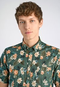 Camicia a maniche corte con bottoni in tessuto verde scuro, caratterizzata da un motivo floreale in arancione, crema e blu teal, con una tasca sul petto e bottoni.