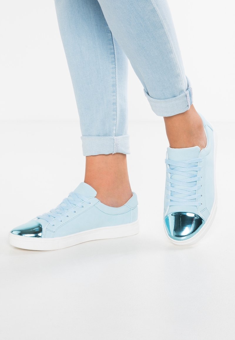 Personne portant des baskets bleu clair avec des embouts métalliques bleus et un jean bleu clair retroussé sur fond blanc.