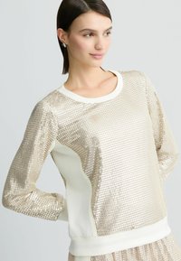 Felpa bronzo con paillettes e un colore avorio per il collo e i dettagli. Presenta maniche lunghe e una superficie testurizzata con una vestibilità aderente.
