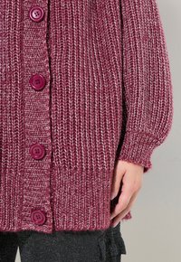 Stickad vinröd kofta med stora lila knappar, ribbad textur och en avslappnad passform, avslutad med en fransad nederkant.