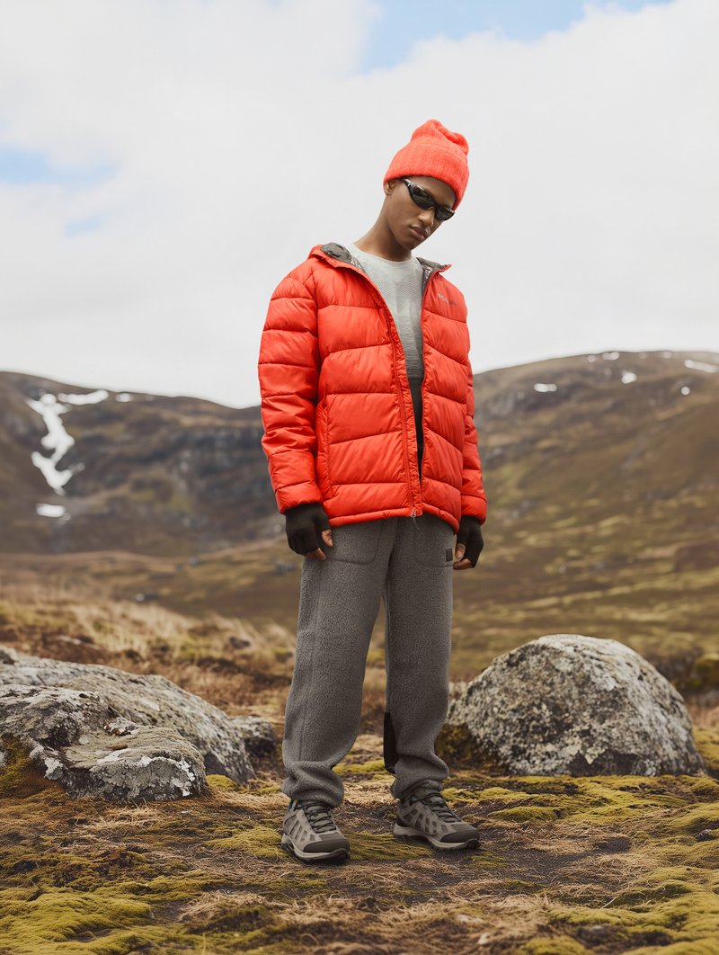 Chaqueta acolchada roja, pantalones de forro polar grises y zapatillas deportivas grises. Los accesorios incluyen un gorro de lana naranja y gafas de sol negras. Entorno al aire libre.