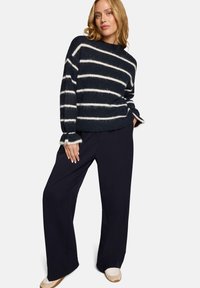 Femme portant un pull en grosse maille à rayures bleu marine et blanc, un pantalon noir à jambes larges, et des baskets blanches, debout avec une main dans la poche.