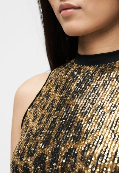 Γυναίκα που φοράει μαύρο και χρυσό τοπ με παγιέτες χωρίς μανίκια, εμφανίζεται από τον ώμο μέχρι το σαγόνι, με ίσια σκούρα μαλλιά.