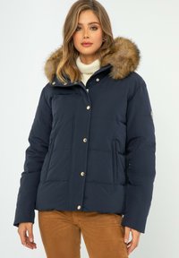 Granatowa kurtka puchowa z kapturem z sztucznego futra, z złotymi zatrzaskami i pikowanym teksturem, w zestawie z kremowym swetrem pod spodem.