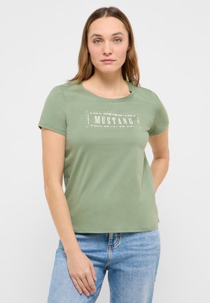 Frau mit hellbraunen Haaren, die ein hellgrünes T-Shirt mit Mustang-Logo und blaue Jeans trägt und mit einer Hand auf der Hüfte steht.