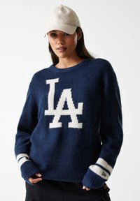Suéter de punto azul marino con un gran logo blanco "LA", con puños a rayas y una textura suave. Combinado con una gorra de color beige claro.