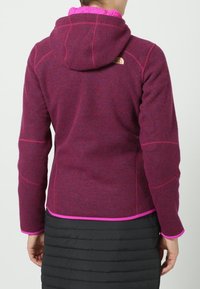 Fioletowa polarowa kurtka z różowym podszyciem kaptura i kontrastującymi różowymi szwami; na plecach znajduje się logo, a krój jest dopasowany.