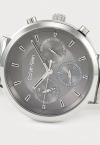 Montre chronographe en métal argenté avec un cadran gris texturé, des indicateurs de date et de jour, et trois sous-dials avec des accents contrastés.