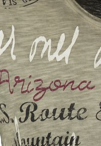 Camiseta verde clara con diversos gráficos de texto en negro, blanco y burdeos, fabricada en un material suave y texturizado, con un escote en forma de barco.