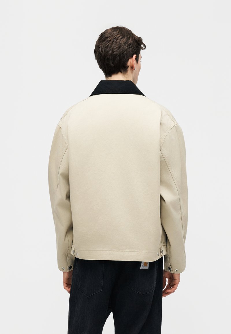 Hombre con chaqueta beige con cuello negro y pantalones negros, de espaldas contra un fondo claro y liso.