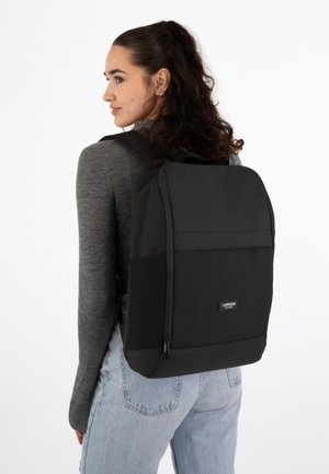 Mochila negra con forma rectangular, hecha de una tela resistente, con un bolsillo delantero con cremallera y una etiqueta de la marca en la parte inferior.
