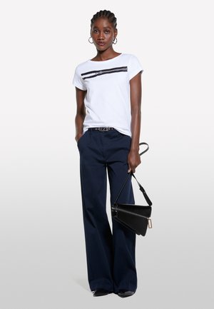 Maglietta bianca con una grafica di cerniera nera, abbinata a pantaloni a gamba larga blu navy. Tiene una borsa nera strutturata con dettaglio di cerniera.