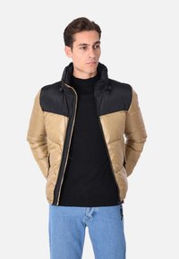 Giacca a vento imbottita con la parte superiore nera e la parte inferiore dorata, realizzata in materiale lucido con sezioni trapuntate, chiusura con zip e cappuccio regolabile.
