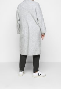 Dlouhý šedý cardigan vyrobený z měkkého materiálu, s uvolněným střihem, spadlými rameny, a kombinovaný se tmavými kalhotami a bílými teniskami.